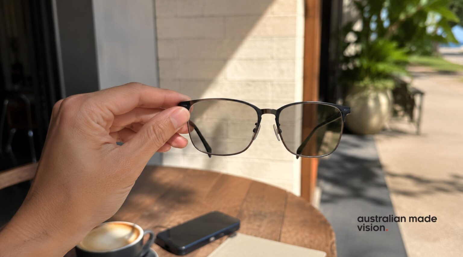 Hand holding a pair of photochromic glasses outdoors, showing lenses darkening automatically when exposed to sunlight.